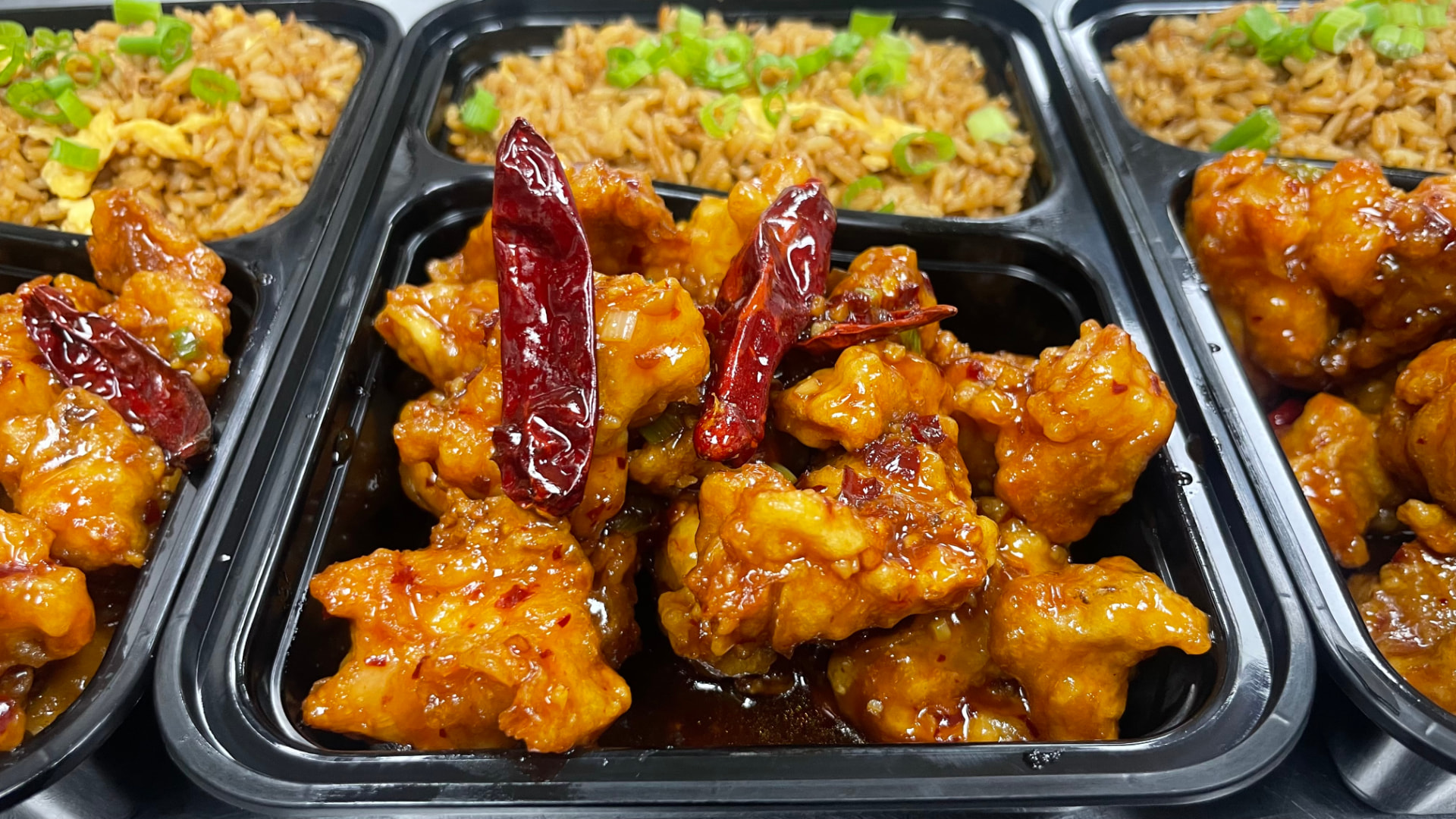 Black takeout containers filled with General Tso's chicken, dried red chilies, and fried rice